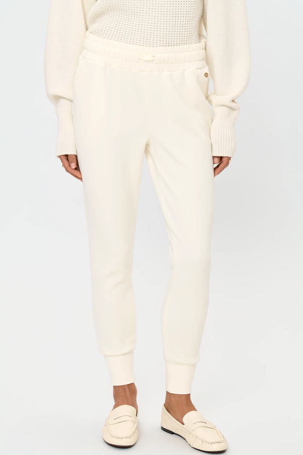 Close-up front view of cream ROMULA stretch pants featuring a comfortable elastic waistband, side pockets, tapered legs, and ribbed ankle cuffs.